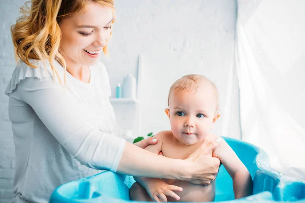 Mãe feliz lavando seu pequeno filho na banheira de plástico do bebê em casa — Fotografia de Stock
