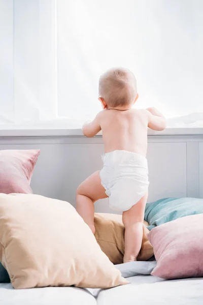 Happy little baby standing on bed with lot of pillows and looking through window — Stock Photo