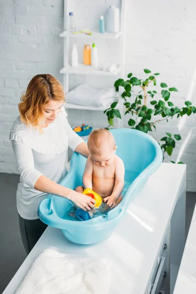 Vista de ángulo alto de la madre lavando a su pequeño niño en la bañera de plástico del bebé en casa - foto de stock