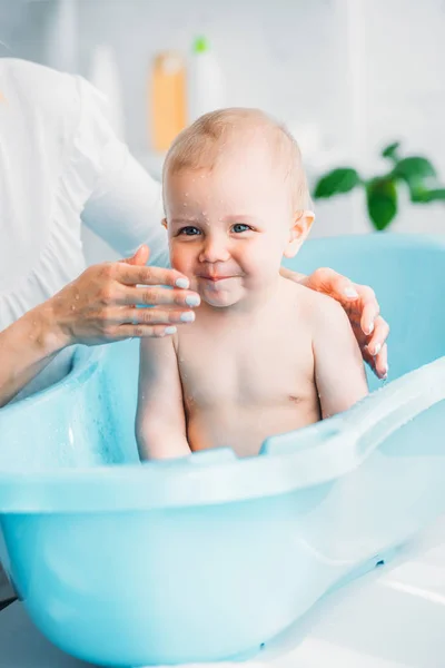 Plan recadré de mère lavant son petit enfant dans une baignoire en plastique pour bébé à la maison — Photo de stock