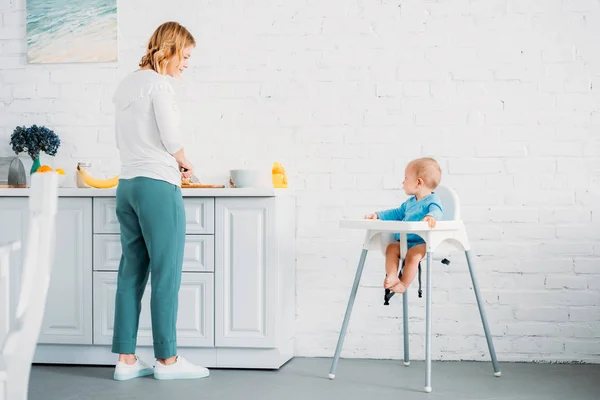 Belle mère préparer le dîner pour son petit enfant alors qu'il est assis dans la chaise d'alimentation à la cuisine — Photo de stock