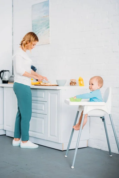 Mother preparing dinner for her little child while he sitting in feeding chair at kitchen — Stock Photo