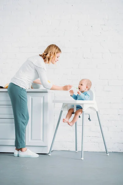 Mère nourrir son petit enfant alors qu'il est assis dans la chaise d'alimentation sur la cuisine à la maison — Photo de stock