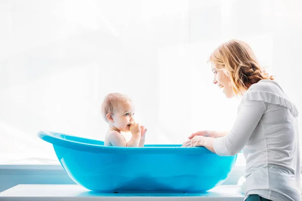 Vista lateral de la madre sonriente lavando a su adorable hijo pequeño en la bañera de plástico del bebé en casa - foto de stock