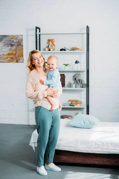 Lachende Mutter trägt ihr kleines Kind und blickt in die Kamera im Schlafzimmer — Stockfoto