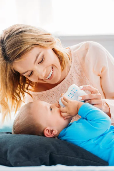 Улыбающаяся мать кормит своего маленького ребенка бутылочкой на кровати дома — стоковое фото