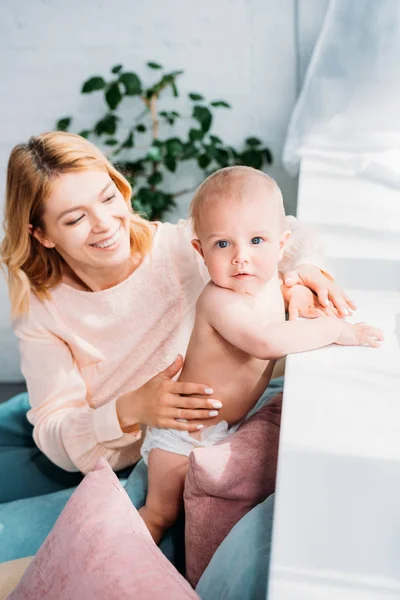 Smiling mother supporting her little child while he leaning on windowsill at home — Stock Photo