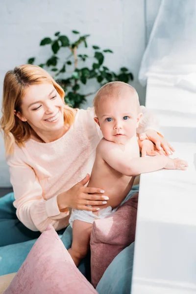 Sonriente madre apoyando a su pequeño hijo mientras él se apoya en el alféizar de la ventana en casa - foto de stock