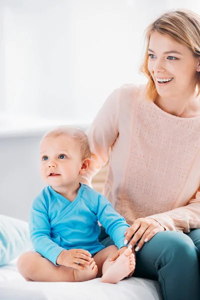 Madre feliz y el niño sentado en la cama en casa y mirando hacia otro lado - foto de stock