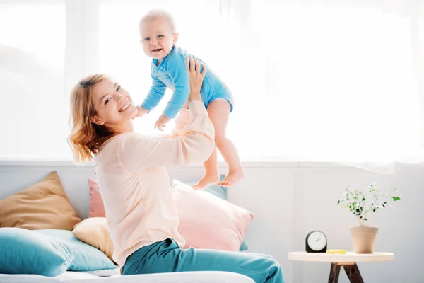 Vue latérale de la mère souriante élevant un petit enfant riant assis sur le lit à la maison — Photo de stock