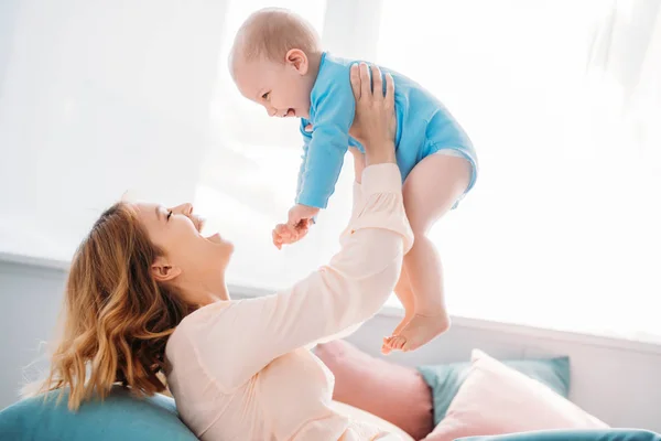 Side view of happy mother raising laughing little child while sitting on bed at home — Stock Photo