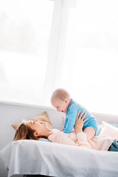 Vue de côté de la mère jouant avec le petit enfant riant tout en étant couché sur le lit à la maison — Photo de stock