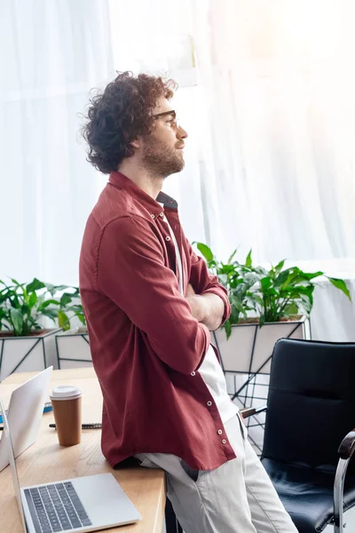 Visão lateral do jovem em óculos de pé com os braços cruzados e olhando para longe no escritório — Fotografia de Stock