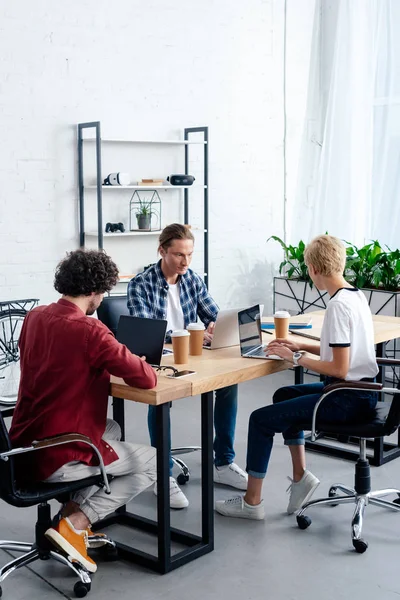 Equipo empresarial joven que utiliza ordenadores portátiles mientras están sentados juntos en el lugar de trabajo - foto de stock