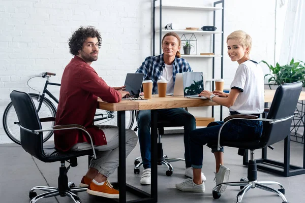 Equipo empresarial joven utilizando computadoras portátiles y mirando a la cámara - foto de stock