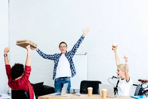 Взволнованная молодая команда стартапов поднимает руки и смотрит на коробки пиццы в офисе — стоковое фото