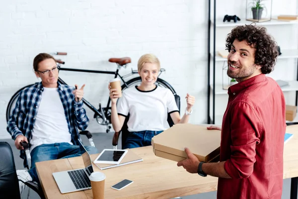 Giovani colleghi sorridenti guardando l'uomo felice che tiene scatole di pizza in ufficio — Foto stock