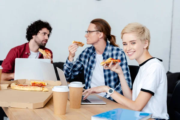 Молода жінка з піцою використовує ноутбук і посміхається на камеру, в той час як чоловіки колеги їдять піцу позаду — стокове фото