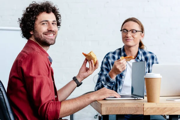 Smiling young men eating pizza and using laptops in office — Stock Photo