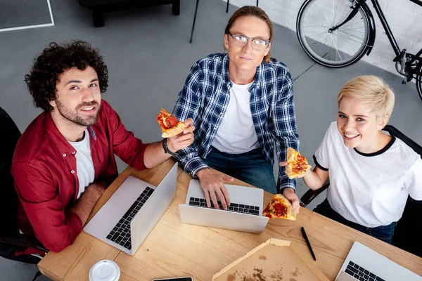 Vue grand angle de sourire jeune équipe de démarrage à l'aide d'ordinateurs portables et de manger de la pizza au bureau — Photo de stock