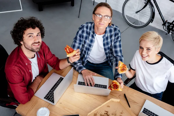 Vista de ángulo alto de sonreír equipo joven puesta en marcha utilizando computadoras portátiles y comer pizza en la oficina - foto de stock