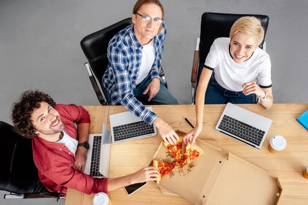 Blick aus der Vogelperspektive auf glückliche junge Kollegen, die am Arbeitsplatz Pizza essen und in die Kamera lächeln — Stockfoto