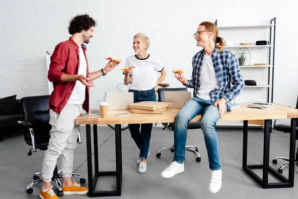 Щасливі молоді колеги їдять піцу і посміхаються один одному в офісі — стокове фото