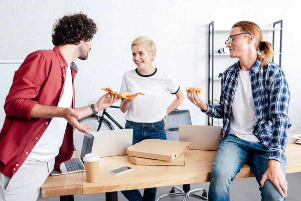 Jóvenes colegas felices sosteniendo pizza y sonriéndose en la oficina - foto de stock