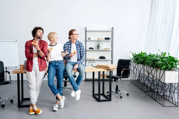 Lächelnde junge Kollegen, die Pizza essen und im Büro wegschauen — Stockfoto