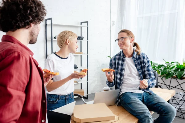 Sonriendo jóvenes colegas comiendo pizza y hablando en la oficina - foto de stock