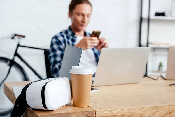 Close-up view of coffee to go, virtual reality headset and man using smartphone behind — Stock Photo