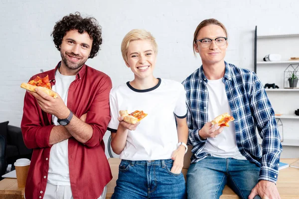 Heureuse jeune équipe de démarrage tenant pizza et souriant à la caméra dans le bureau — Photo de stock