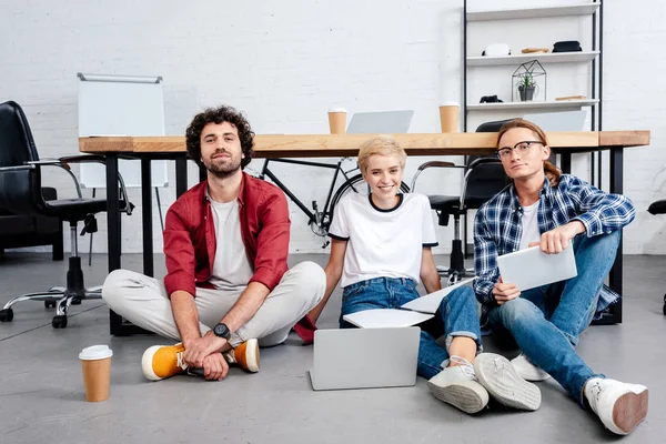 Sonriente joven equipo de puesta en marcha sentado en el suelo y trabajando juntos - foto de stock