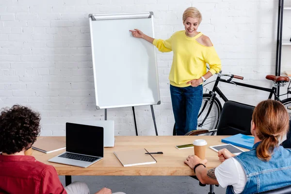 Lächelnde junge Frau zeigt auf leeres Whiteboard und blickt auf männliche Kollegen im Büro — Stockfoto