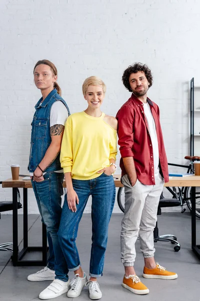 Jóvenes colegas de negocios de pie juntos y sonriendo a la cámara en la oficina - foto de stock