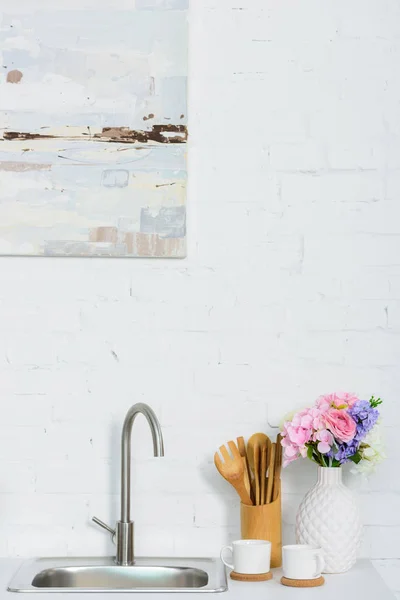 Florero con flores de colores, tazas y utensilio de madera en el mostrador de la cocina cerca del fregadero - foto de stock