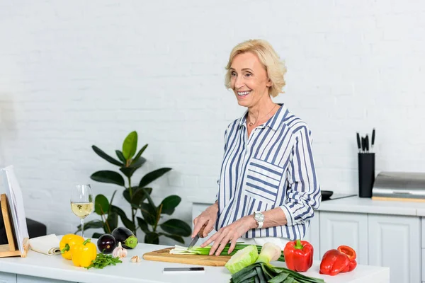 Sorrindo atraente mulher de cabelo cinza cortando cebola verde na cozinha — Fotografia de Stock