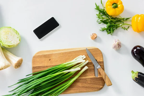 Green onion on wooden board, smartphone with blank screen on white tabletop in kitchen — Stock Photo