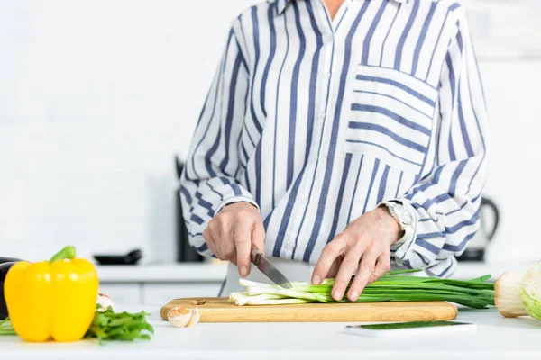 Image recadrée d'une femme aux cheveux gris coupant l'oignon vert sur une planche en bois dans la cuisine — Photo de stock