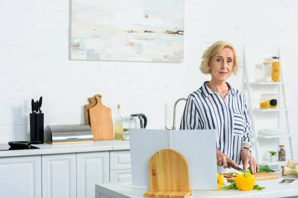 Attraktive grauhaarige Frau schneidet grüne Zwiebel in der Küche und schaut in die Kamera — Stockfoto
