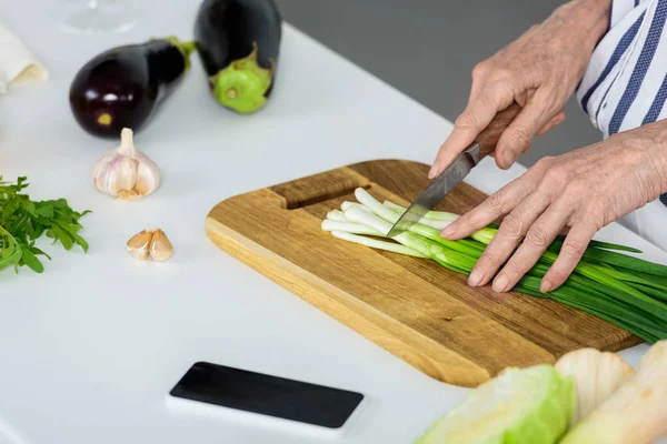 Imagem cortada de mulher de cabelo cinza cortando cebola verde na placa de madeira na cozinha — Fotografia de Stock