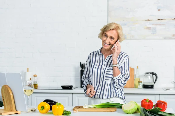 Sonriendo atractiva mujer de pelo gris cortando cebolla verde en la cocina y hablando por teléfono inteligente - foto de stock