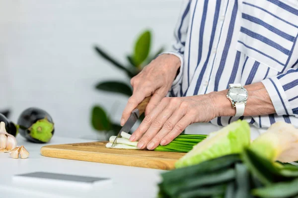 Abgeschnittenes Bild einer grauhaarigen Frau, die in der Küche frische grüne Zwiebeln auf Holzbrettern schneidet — Stockfoto