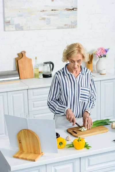 High angle view of attractive grey hair woman cutting green onion in kitchen — Stock Photo