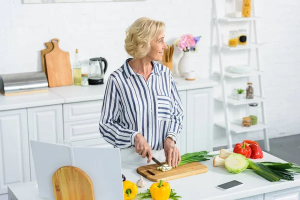 Привлекательные седые волосы женщина режет зеленый лук на кухне и смотрит в сторону — стоковое фото