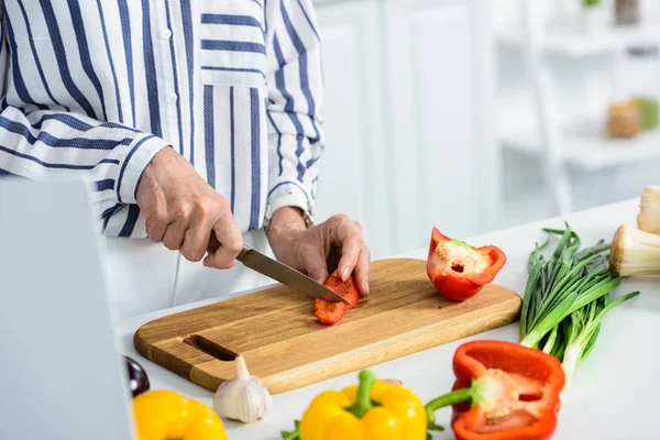 Cropped image of grey hair woman cutting red bell pepper on cutting board in kitchen — Stock Photo