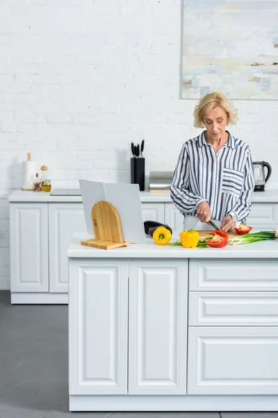 Atractiva mujer de pelo gris cocinando en cocina y cortando verduras - foto de stock