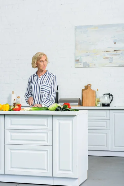 Bella donna capelli grigi tagliare verdure in cucina e distogliere lo sguardo — Foto stock