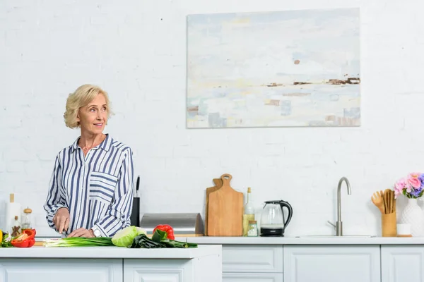 Attraente donna capelli grigi tagliare verdure in cucina e distogliere lo sguardo — Foto stock