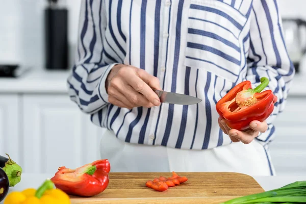 Abgeschnittenes Bild einer grauhaarigen Frau, die in der Küche rote Paprika auf einem Holzbrett schneidet — Stockfoto